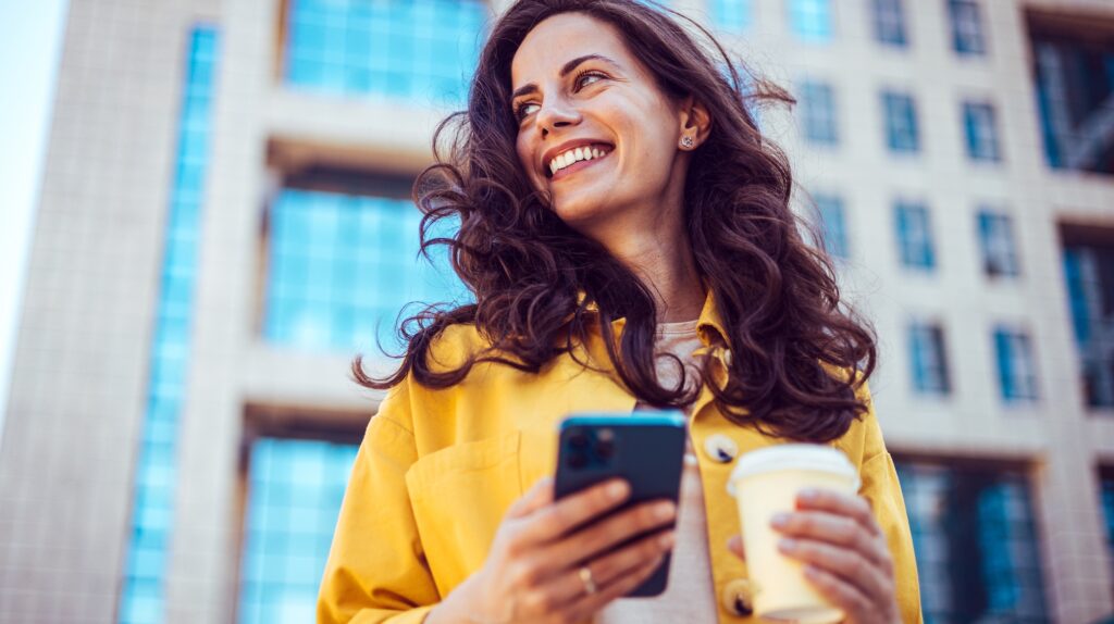 Smiling woman holding coffee cup while accessing curriculum management platform on mobile device.