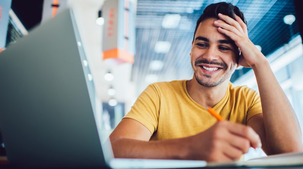 Young smiling man using his laptop to browse university courses on his laptop