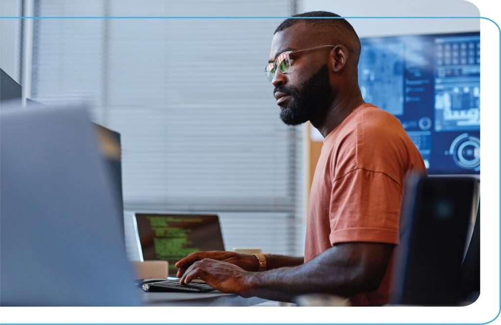 Young black man sitting at a desk surrounded by tech and monitors.